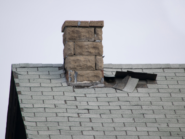 Storm-damaged shingles lifted away from the roof deck beside a brick chimney